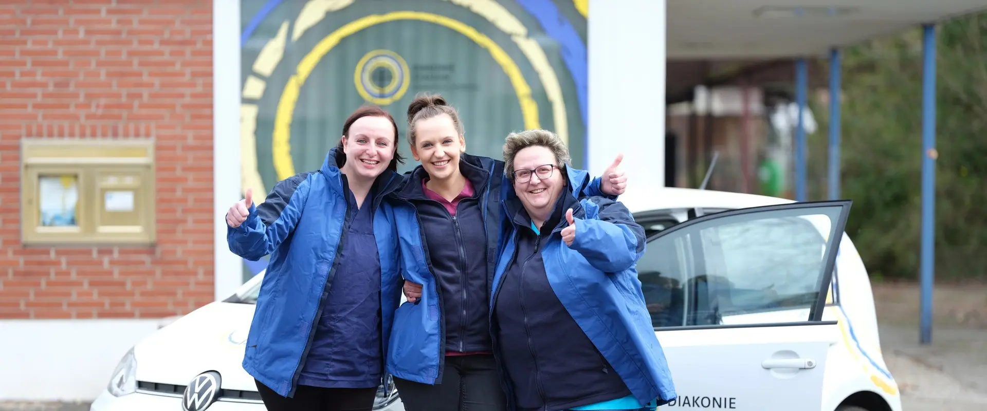 Fröhliches Pflegeteam der ambulanten Pflege steht am Auto und lacht in die Kamera