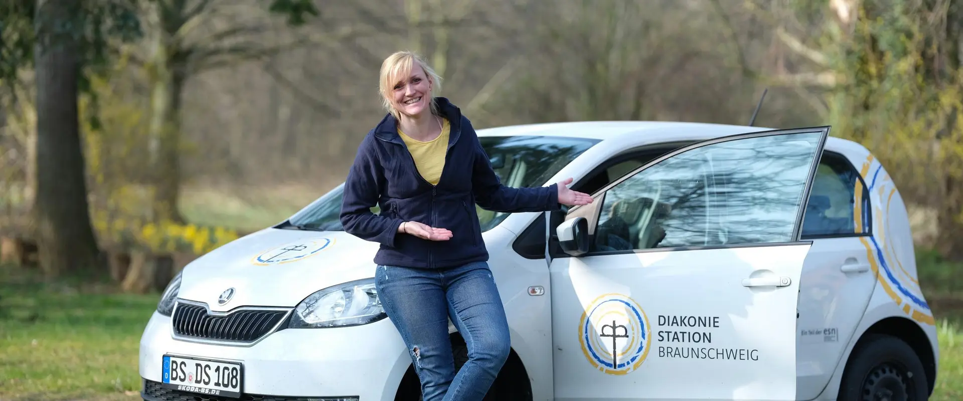 Ambulante Pflegefachkraft steht an einem Auto der Diakoniestation und lacht in die Kamera
